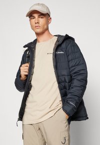 Marineblauwe pufferjack met capuchon, voorzien van een ritssluiting en Columbia-logo. Beige t-shirt eronder, gecombineerd met lichtbeige cargo-broeken.