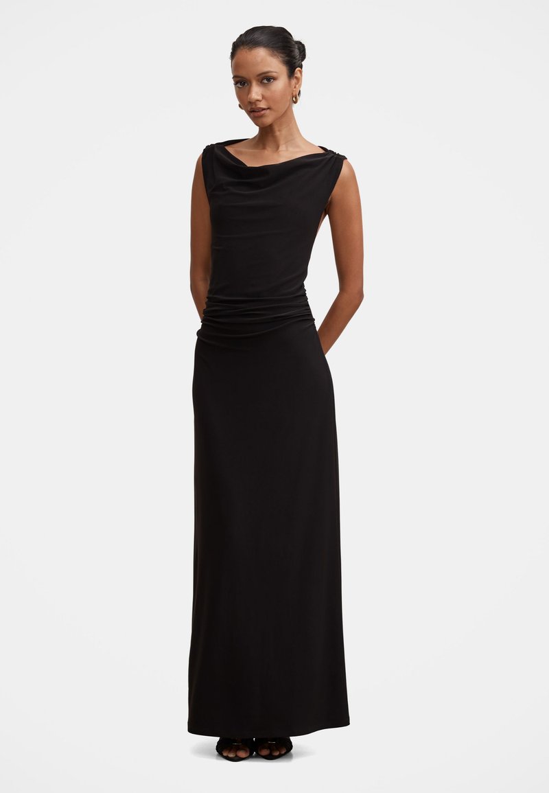 Femme portant une robe noire longue sans manches avec un décolleté drapé, debout devant un fond blanc.