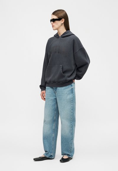 Femme portant un sweat à capuche noir oversize, un jean large bleu clair, des ballerines noires, des lunettes de soleil noires et des boucles d'oreilles argentées, debout une main dans la poche.