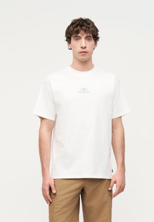 Νέος άνδρας με κοντά σγουρά μαλλιά, φορώντας λευκό κοντομάνικο μπλουζάκι με μικρό λογότυπο και καφέ cargo παντελόνια, στέκεται μπροστά σε απλό φόντο.