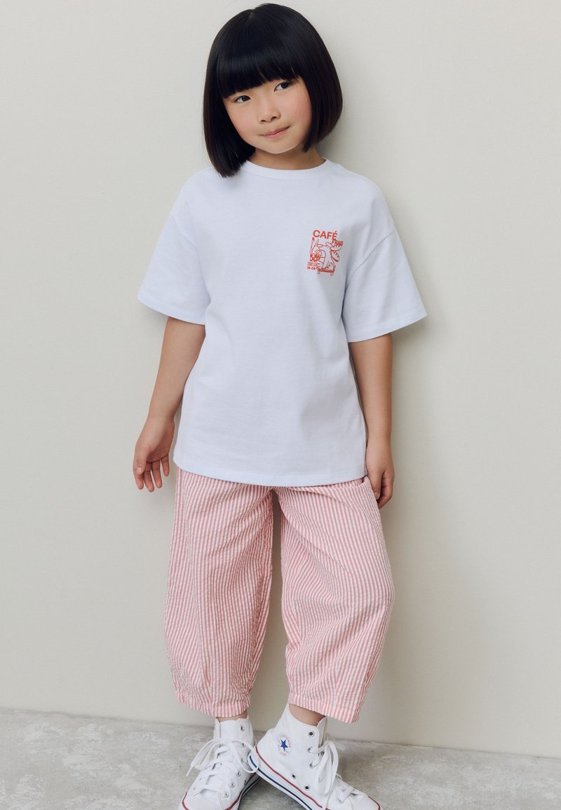 T-shirt en coton blanc avec un imprimé graphique rouge sur la poitrine gauche, associée à un pantalon court rayé rouge et blanc et des chaussures montantes blanches.