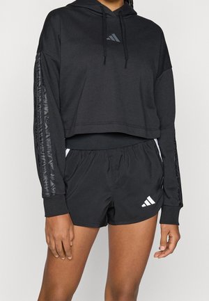 Personne portant un sweat à capuche court noir Adidas et un short de sport noir avec le logo Adidas, debout devant un fond uni.