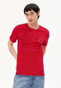 T-shirt rouge à manches courtes en coton, avec un petit graphisme circulaire sur la poitrine. Texture lisse avec un col rond.