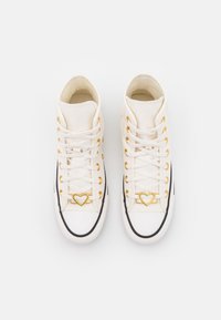 Białe trampki za kostkę z płótna, z złotymi oczkami i sprzętem w kształcie serca, z czarną gumową podeszwą i sznurówkami.