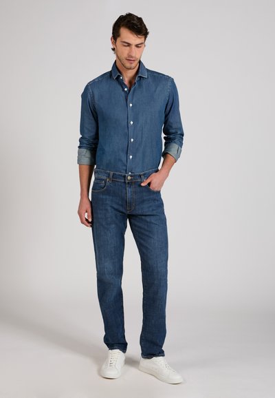 Man wearing blue denim shirt and jeans with white sneakers, standing with one hand in pocket against plain background.