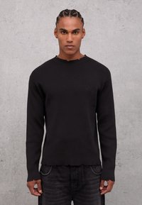 Top lungo a maniche nere a coste con scollatura a girocollo, caratterizzato da un orlo grezzo e un sottile logo sul petto. Tessuto testurizzato.