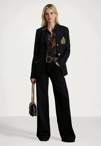 Szőke nő fekete blézert visel arany címerrel, mintás blúzt, széles fekete nadrágot, fekete övet, és egy fekete táskát tart arany lánccal.