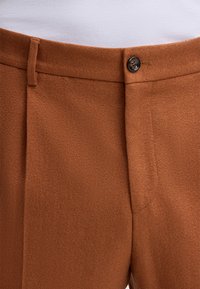 Bruine broek van gevlochten stof, voorzien van een enkele knoopsluiting, diagonale zakken en een getailleerde pasvorm bij de taille.