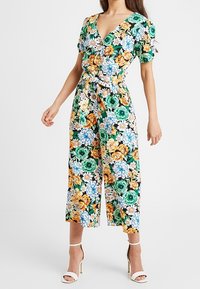 Kwiatowy kombinezon w odcieniach zieleni, z dużymi pomarańczowymi, niebieskimi i białymi kwiatami. Krótkie bufiaste rękawy, dekolt w kształcie litery V, przewiązany w pasie i szerokie, skrócone nogawki.