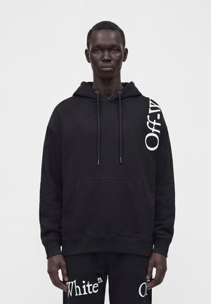 Black hoodie with white "Off-White" text on the left shoulder, worn by a standing person with their hands relaxed by their sides.