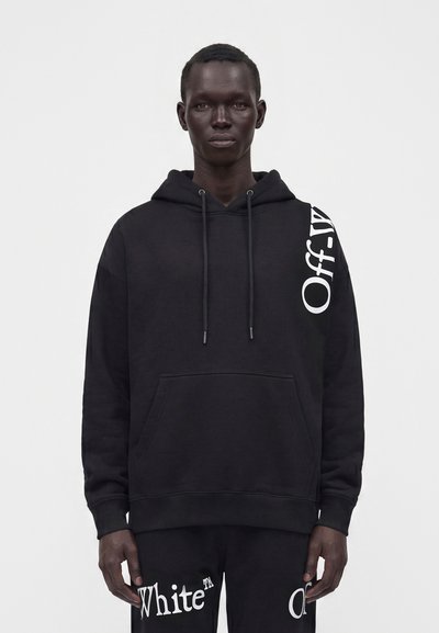 Μαύρο φούτερ με κουκούλα με λευκό κείμενο "Off-White" στον αριστερό ώμο, φορεμένο από ένα όρθιο άτομο με τα χέρια χαλαρά στα πλάγια.