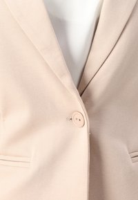 Ljust beige blazer med slät tyg, notch-öppningar, en enkel knäppning och två framfickor.