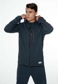 Hoodie zippé bleu marine foncé en tissu doux, présentant une poche poitrine zippée et un patch de marque discret. Texture lisse, coupe décontractée.