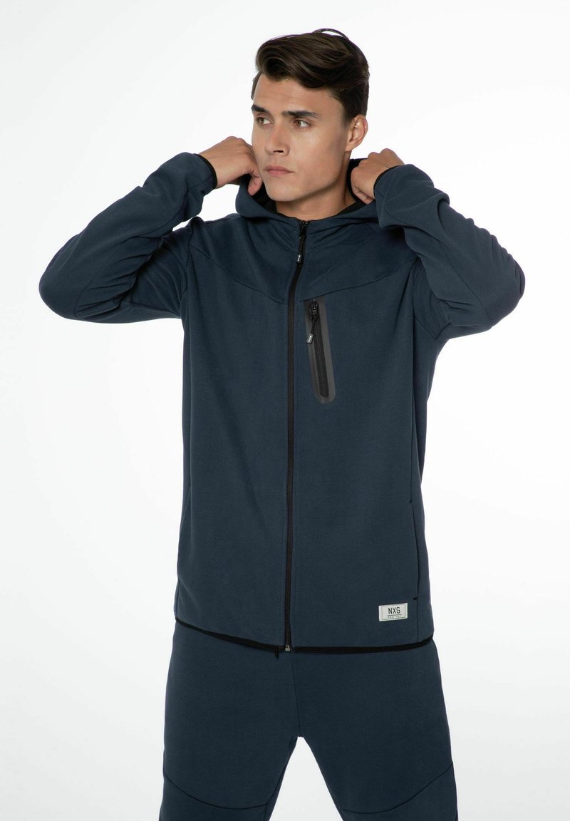 Hoodie zippé bleu marine foncé en tissu doux, présentant une poche poitrine zippée et un patch de marque discret. Texture lisse, coupe décontractée.