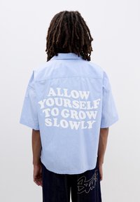 Camicia a maniche corte a righe blu con una vestibilità rilassata, caratterizzata dal testo "PERMETTI AL TUO IO DI CRESCERE LENTAMENTE" stampato in grassetto bianco sulla parte posteriore.