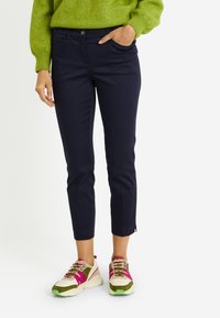 Persona vistiendo pantalones azul marino ajustados hasta el tobillo, zapatillas gruesas verdes y rosas, y un suéter de punto verde con una mano en el bolsillo.