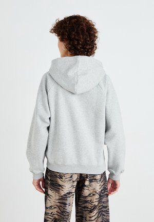 Personne aux cheveux courts et bouclés portant un sweat à capuche gris clair et un pantalon marron à motif tigre, vue de dos sur un fond blanc uni.