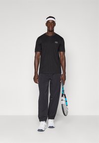 Črna športna majica s kratkimi rokavi, logo na prsih, v kombinaciji s črnimi športnimi hlačami in belimi športnimi čevlji, drži tenis lopar.