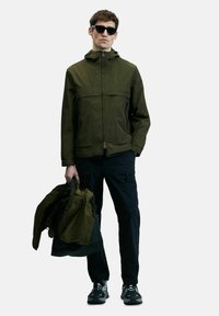 Veste imperméable vert olive avec une fermeture éclair à l'avant, capuche réglable et deux poches latérales, associée à un pantalon cargo noir et des baskets.