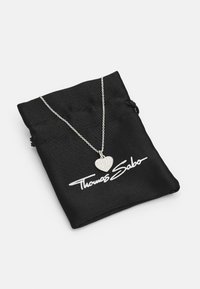 Collar de corazón de plata sobre una cadena delgada, colocado sobre una bolsa de tela negra con el logo "Thomas Sabo" impreso en blanco.