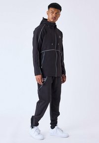 Chándal deportivo negro que incluye una sudadera con cremallera y pantalones a juego. Los acentos en gris claro resaltan las costuras y los bolsillos. Zapatillas blancas completan el look.