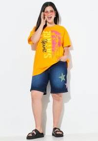 Camiseta naranja oversized con estampado gráfico, combinada con pantalones cortos de mezclilla oscuros con acentos de estrellas. Sandalias negras con suela gruesa.