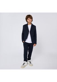 Marinblå kostymjacka med enkla knappar och slim fit, matchad med byxor i samma tyg och vita sneakers. Slät tyg, minimal design.