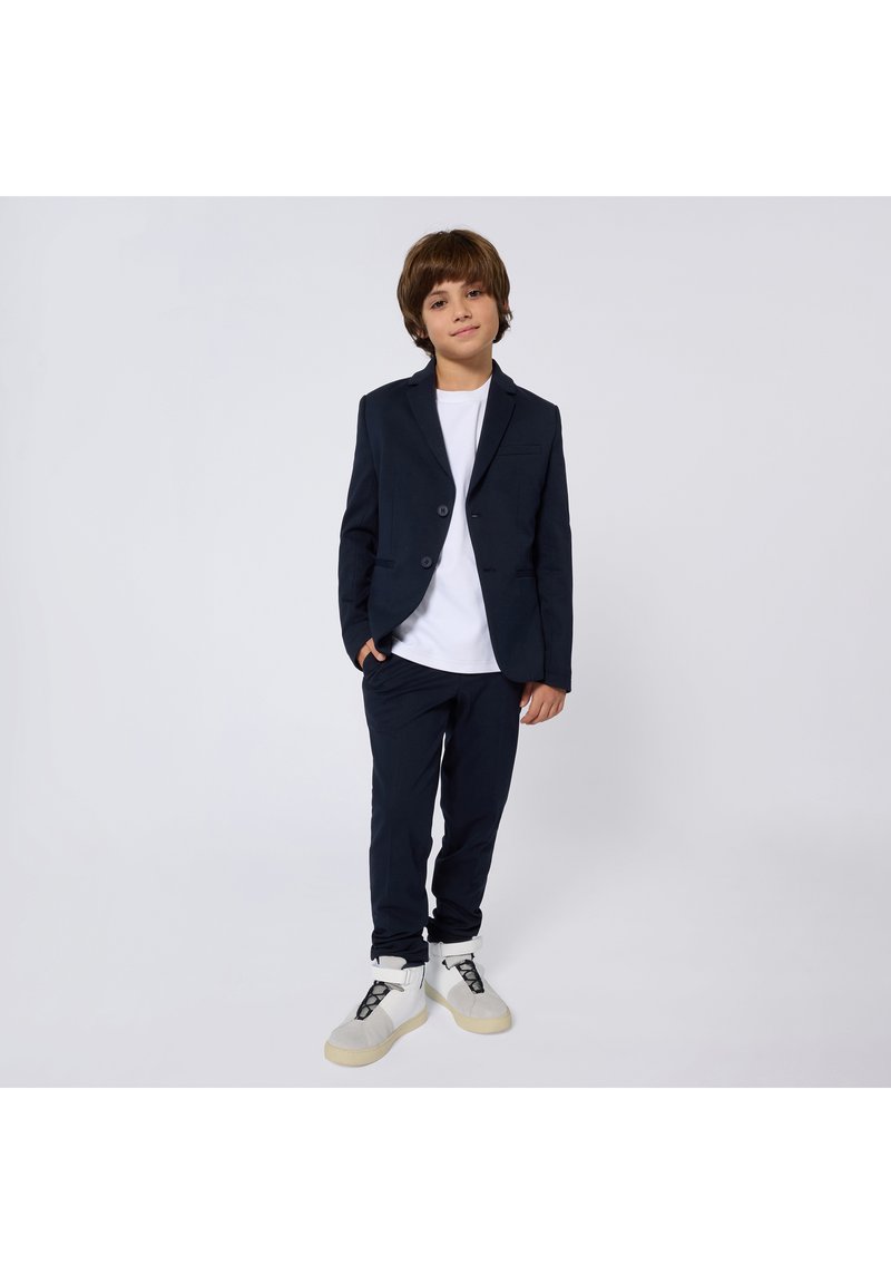 Marinblå kostymjacka med enkla knappar och slim fit, matchad med byxor i samma tyg och vita sneakers. Slät tyg, minimal design.