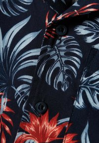 Tessuto blu navy con motivo floreale colorato in rosso e azzurro chiaro; presenta un bottone nero e un dettaglio colletto.