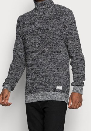 Homme portant un pull à col roulé tricoté chiné noir et blanc avec manches longues et un pantalon noir, debout devant un fond uni.