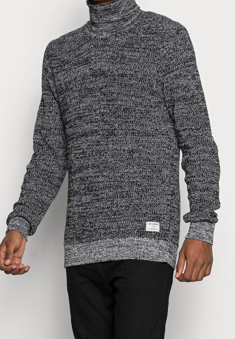 Homme portant un pull à col roulé tricoté chiné noir et blanc avec manches longues et un pantalon noir, debout devant un fond uni.