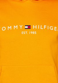 Žltá mikina s kapucňou a predným vreckom, s textom "TOMMY HILFIGER" a červenobielo-modrým logom. Mäkkučký bavlnený materiál.