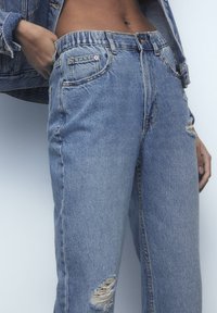 Vaqueros de mezclilla azul claro con corte relajado, cintura alta y detalles desgastados en las rodillas. Accesorios de hardware mínimos y detalles en los bolsillos.