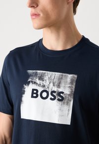 T-shirt in cotone blu navy con una grafica rettangolare bianca che presenta il testo in nero "BOSS" e uno sfondo testurizzato. Collo rotondo.