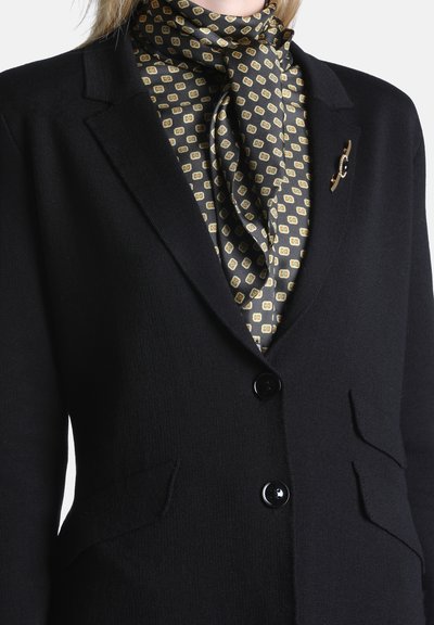 Blazer noir à deux boutons par-dessus une écharpe noire et dorée à motifs nouée au cou, avec une broche dorée sur le revers.