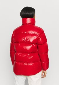 Henkilö, jolla on lyhyet mustat hiukset, pukeutuneena kiiltävään punaiseen untuvatakkiin ja valkoisiin housuihin, kääntynyt pois yksinkertaista vaaleaa taustaa vasten.