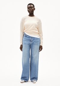 Licht perzikkleurige sweat met lange mouwen, gecombineerd met lichtblauwe jeans met wijde pijpen en zilveren schoenen. De outfit heeft een relaxed fit en een zachte textuur.