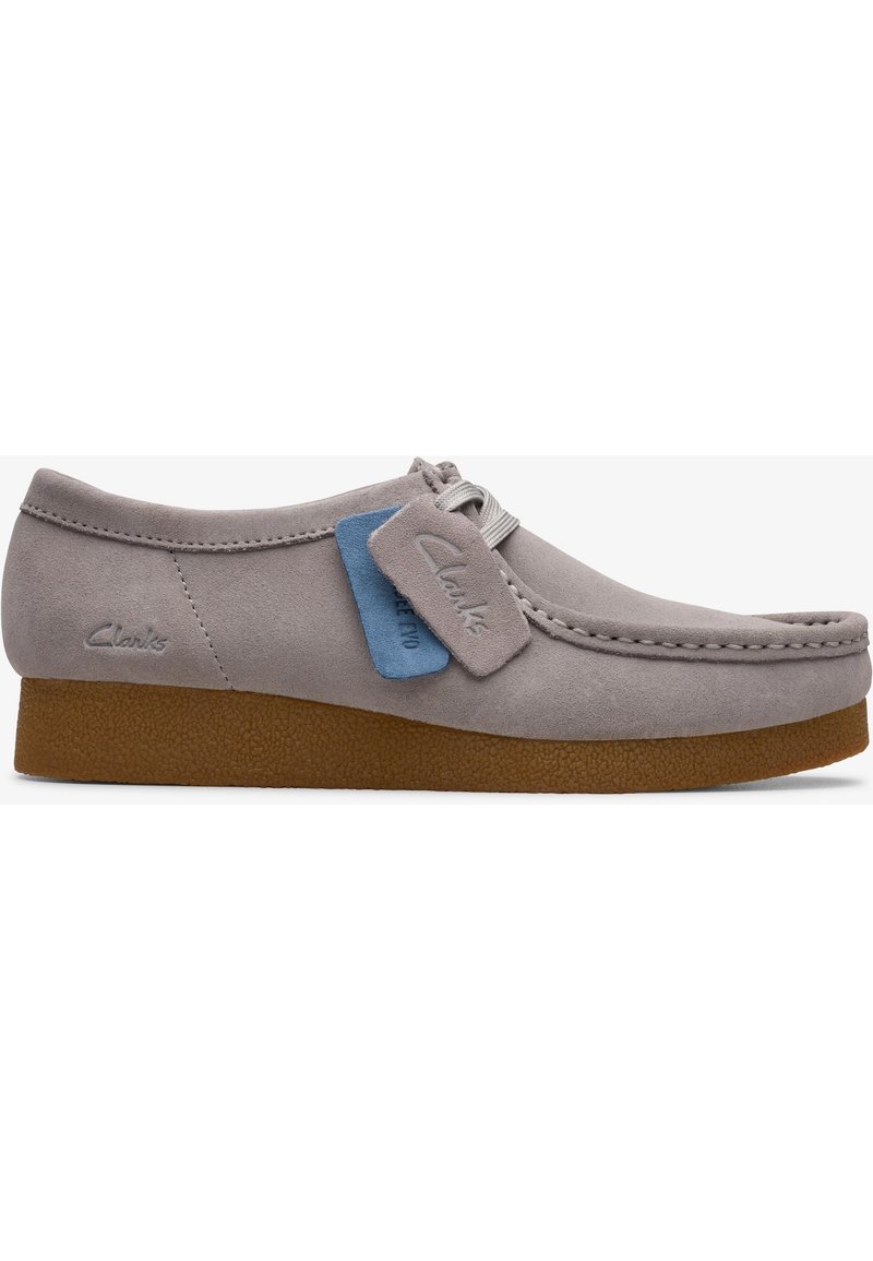 Zapato de gamuza gris con puntera redonda y suela de goma. Presenta un cordón decorativo y una etiqueta azul con el logo de "Clarks".