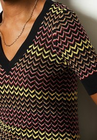 Neulepaita, jossa on V-kaula-aukko, lyhyet hihat ja siksak-kuvio punaisissa, mustissa, keltaisissa ja kultaisissa sävyissä. Rakenteellinen lanka antaa syvyyttä.