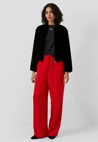 Schwarze Samt-Zip-Up-Bomberjacke über einem schwarzen Strickoberteil, kombiniert mit hochtaillierten, lässig geschnittenen roten Hosen. Schwarze Schuhe runden das Outfit ab.