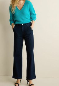 Pull en tricot turquoise à col en V avec motif texturé associé à un pantalon large bleu foncé. Montre en argent minimaliste et talons aiguilles noirs à lanières.