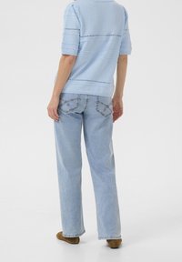 Persona vestida con un top de punto azul claro y jeans sueltos de lavado claro, de pie de espaldas a la cámara sobre un fondo blanco plano.