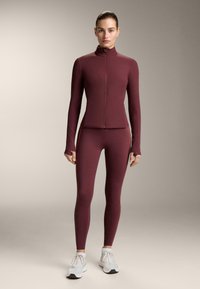 Sportska jakna i tajice boje tamno crvene, obje s visokim ovratnikom i potpunim patentnim zatvaračem, izrađene od glatkog i rastezljivog materijala, nošene uz bijele tenisice.
