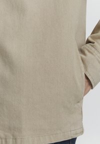 Beige teksturert jakke med diagonalt ripemønster; har en avslappet passform og en lomme med innfelt detalj på siden.