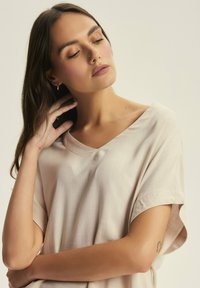 Beżowa bluzka z krótkim rękawem i dekoltem w serek, wykonana z miękkiego materiału o delikatnym połysku. Posiada luźny krój oraz subtelne detale w szwie.