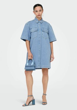 Lichtblauwe jeanshemdjurk. Heeft korte mouwen, knoopsluiting aan de voorkant en twee borstzakken. Gepaard met een blauwe handtas en sleehakken.