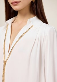 Blusa bianca a maniche lunghe con colletto, caratterizzata da pieghe verticali e un dettaglio in catena dorata che scende sul davanti. Tessuto liscio e leggero.