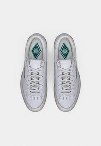 White leather sneakers with a rounded toe, textured laces, perforated detailing, and green accents on the insole and side logo.