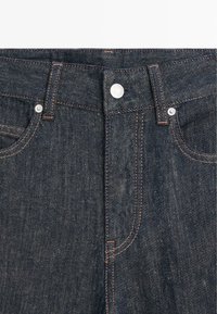 Dunkelblaue Denim-Jeans in Nahaufnahme, die Knopf, Gürtelschlaufen und Fronttaschen mit orangefarbener Naht zeigen.