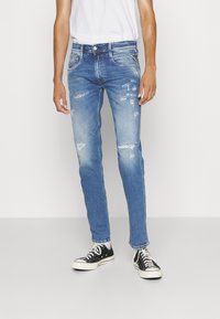 Light blue distressed denim jeans with a slim fit, featuring frayed details and a classic five-pocket design, paired with black sneakers.